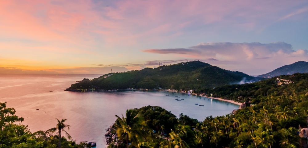 Panoramic view of a tropical bay at sunset, with lush green hillsides dotted with palm trees and a village along the coastline, under a pink and orange sky.