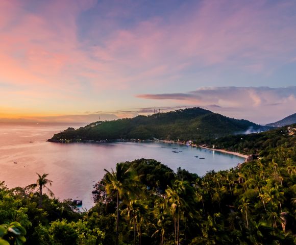 Panoramic view of a tropical bay at sunset, with lush green hillsides dotted with palm trees and a village along the coastline, under a pink and orange sky.