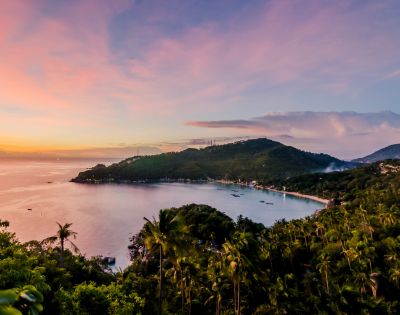 Panoramic view of a tropical bay at sunset, with lush green hillsides dotted with palm trees and a village along the coastline, under a pink and orange sky.