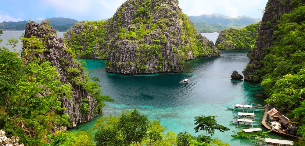 Tropical limestone karst islands covered in lush green vegetation rise from clear turquoise waters where several small boats are moored near a cluster of stilt houses.