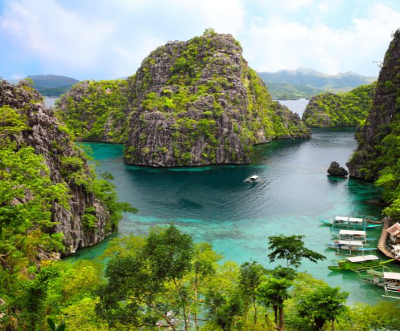 Tropical limestone karst islands covered in lush green vegetation rise from clear turquoise waters where several small boats are moored near a cluster of stilt houses.