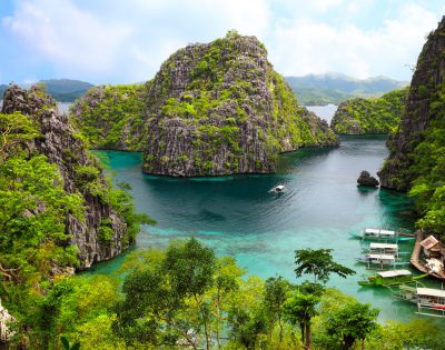 Tropical limestone karst islands covered in lush green vegetation rise from clear turquoise waters where several small boats are moored near a cluster of stilt houses.
