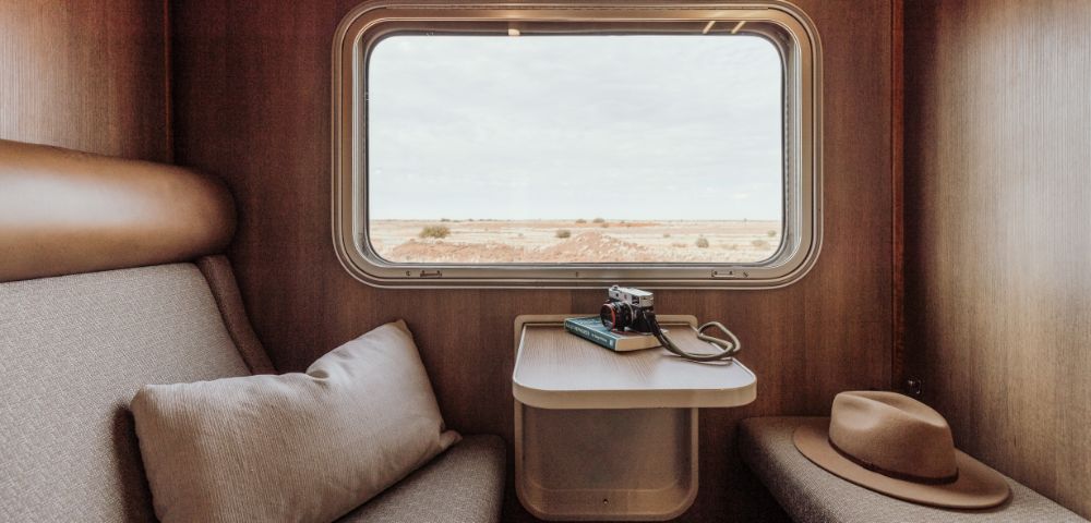Cozy train cabin interior with a large window revealing a vast desert landscape. A camera and book lie on the table, while a hat rests on a seat, suggesting travel.
