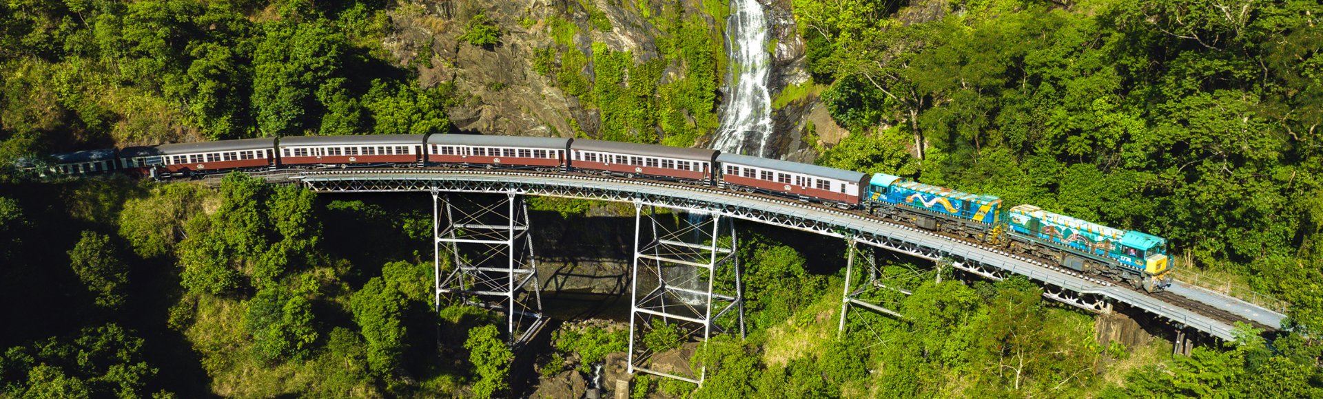 A train crosses a metal bridge over lush greenery, with a cascading waterfall in the background, conveying a sense of adventure and natural beauty.