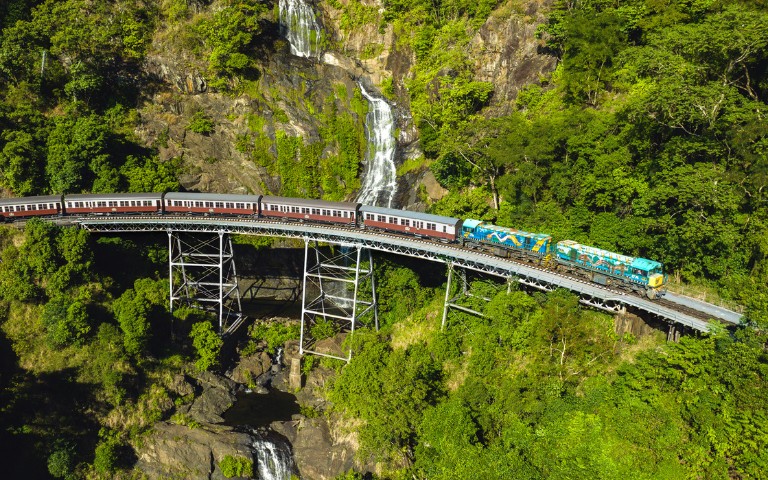 A train crosses a metal bridge over lush greenery, with a cascading waterfall in the background, conveying a sense of adventure and natural beauty.
