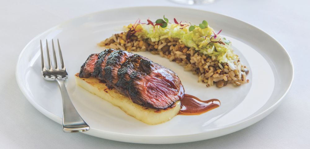 Grilled steak on creamy potato puree with a demi-glace sauce, paired with mixed rice topped with shredded greens, on a white plate, elegant setting.