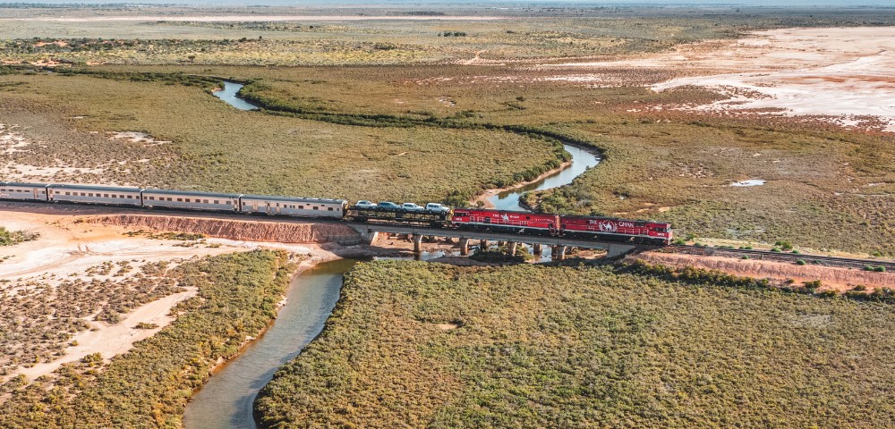Aerial view of a red train crossing a bridge over a winding river in a vast, arid landscape. The scene conveys a sense of motion and isolation.