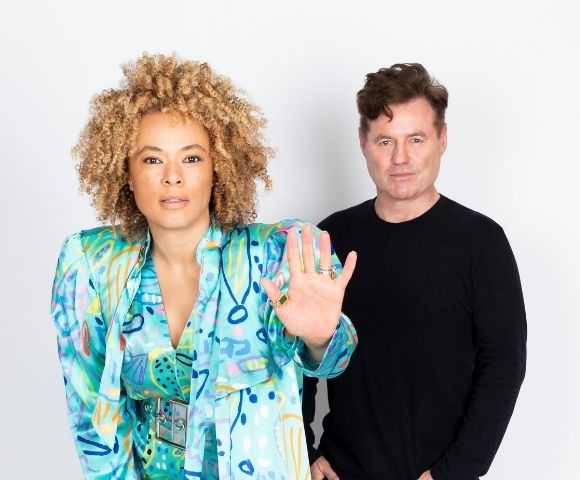 A woman with curly blonde hair wearing a colorful patterned jacket and a man in a black shirt stand against a white background, with the woman holding up her hand.