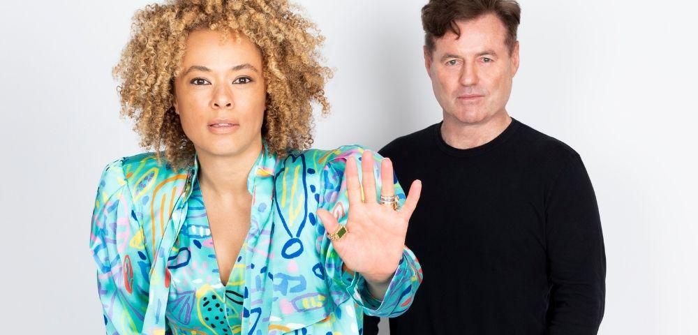 A woman with curly blonde hair wearing a colorful patterned jacket and a man in a black shirt stand against a white background, with the woman holding up her hand.
