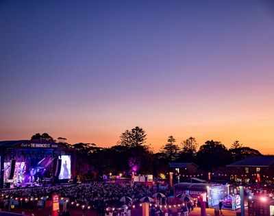 An outdoor concert at dusk with a large crowd gathered in front of a brightly lit stage.