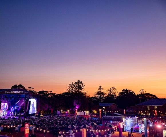 An outdoor concert at dusk with a large crowd gathered in front of a brightly lit stage.