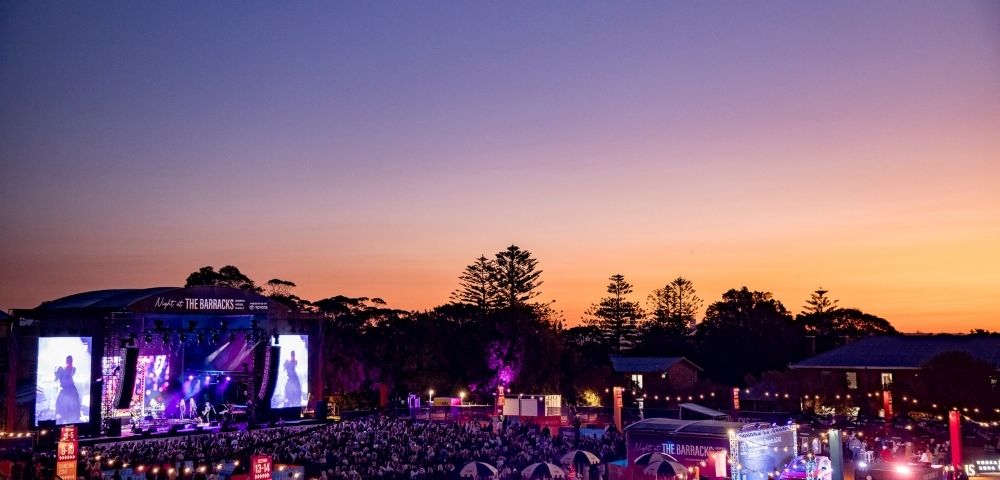 An outdoor concert at dusk with a large crowd gathered in front of a brightly lit stage.
