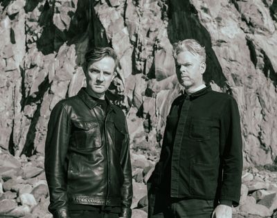 Two men stand in front of a rocky cliff face. The man on the left wears a leather jacket and the man on the right wears a dark collared shirt.