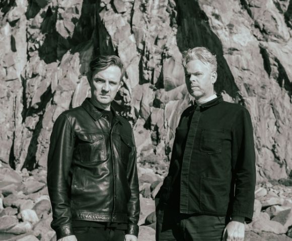 Two men stand in front of a rocky cliff face. The man on the left wears a leather jacket and the man on the right wears a dark collared shirt.