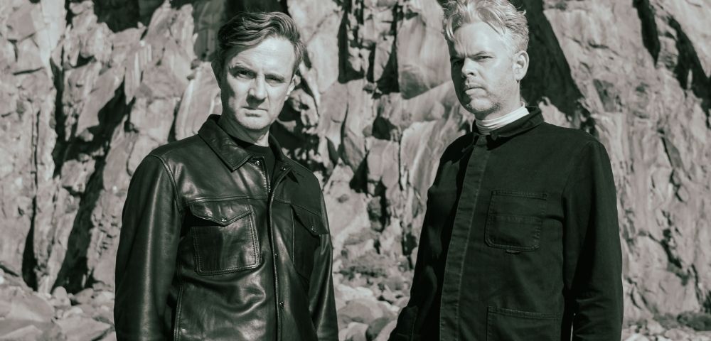 Two men stand in front of a rocky cliff face. The man on the left wears a leather jacket and the man on the right wears a dark collared shirt.