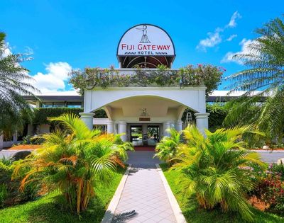 The entrance to the Fiji Gateway Hotel with a paved walkway leading to the building, flanked by lush green palm trees and tropical foliage under a bright blue sky.