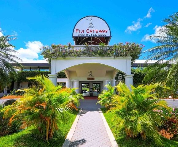 The entrance to the Fiji Gateway Hotel with a paved walkway leading to the building, flanked by lush green palm trees and tropical foliage under a bright blue sky.