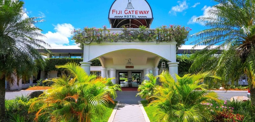 The entrance to the Fiji Gateway Hotel with a paved walkway leading to the building, flanked by lush green palm trees and tropical foliage under a bright blue sky.
