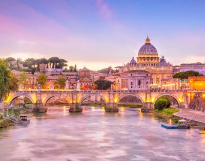 Scenic view of Rome at sunset, featuring the lit-up St. Peter's Basilica and a historic stone bridge over the Tiber River, creating a warm, serene ambiance.