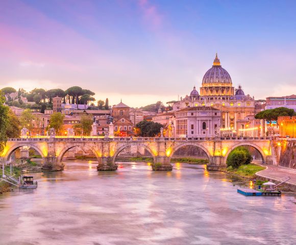 Scenic view of Rome at sunset, featuring the lit-up St. Peter's Basilica and a historic stone bridge over the Tiber River, creating a warm, serene ambiance.
