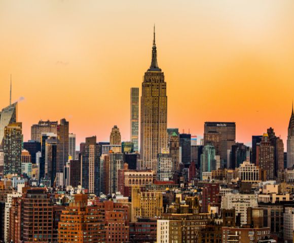 Sunset skyline of a city with tall skyscrapers, including a prominent central building. The sky glows with warm orange hues, creating a tranquil atmosphere.