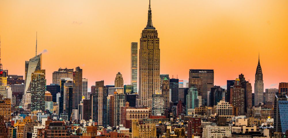 Sunset skyline of a city with tall skyscrapers, including a prominent central building. The sky glows with warm orange hues, creating a tranquil atmosphere.