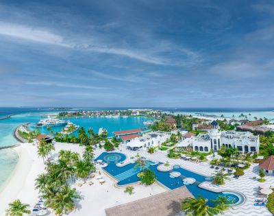 Aerial view of a tropical resort with vibrant blue pools, white buildings, and palm trees. The serene ocean and boats are visible in the background.