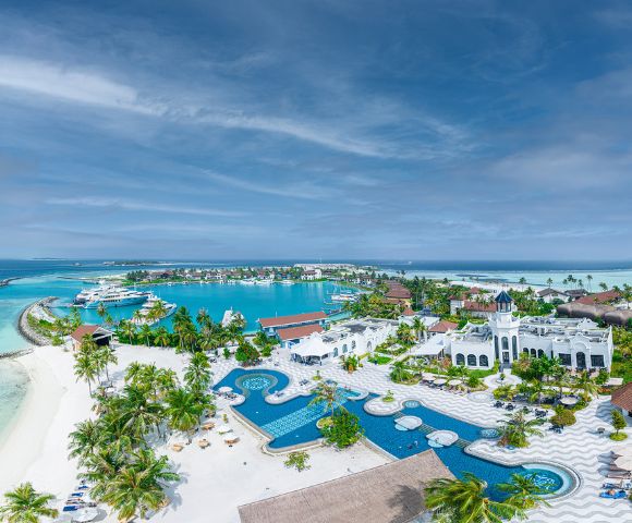 Aerial view of a tropical resort with vibrant blue pools, white buildings, and palm trees. The serene ocean and boats are visible in the background.