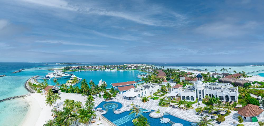 Aerial view of a tropical resort with vibrant blue pools, white buildings, and palm trees. The serene ocean and boats are visible in the background.