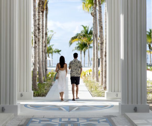 A couple walks away between tall white columns and palm trees toward a sunny beach, conveying serenity and tropical relaxation.