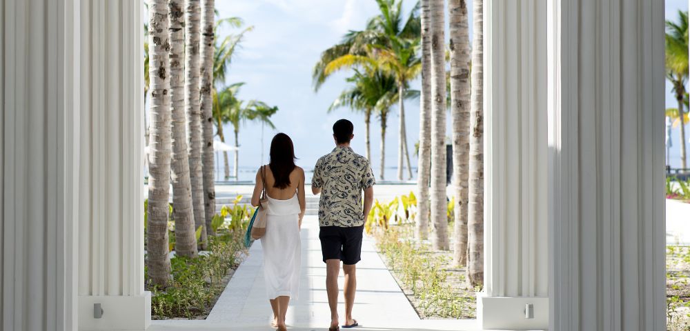 A couple walks away between tall white columns and palm trees toward a sunny beach, conveying serenity and tropical relaxation.