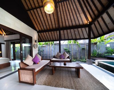 Outdoor living area with wooden furniture, purple and cream pillows, a coffee table, and a thatched roof, next to a swimming pool.