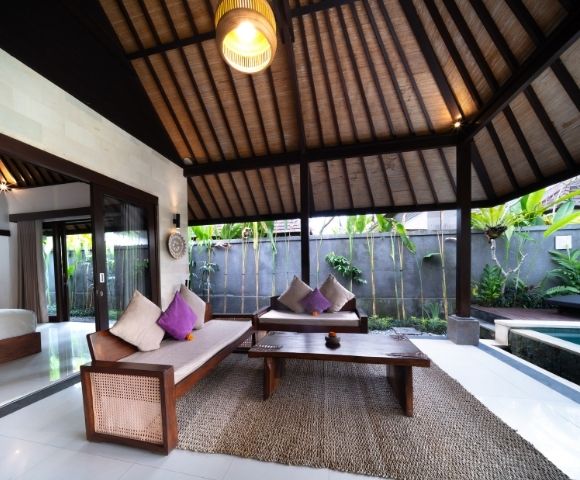 Outdoor living area with wooden furniture, purple and cream pillows, a coffee table, and a thatched roof, next to a swimming pool.
