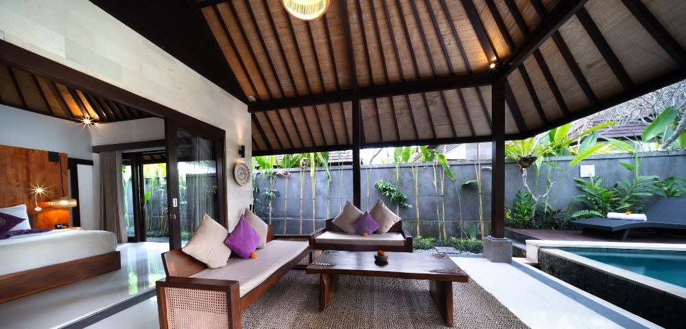 Outdoor living area with wooden furniture, purple and cream pillows, a coffee table, and a thatched roof, next to a swimming pool.