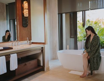 Woman in a green robe sitting on the edge of a white bathtub in a modern bathroom with large windows overlooking lush greenery.