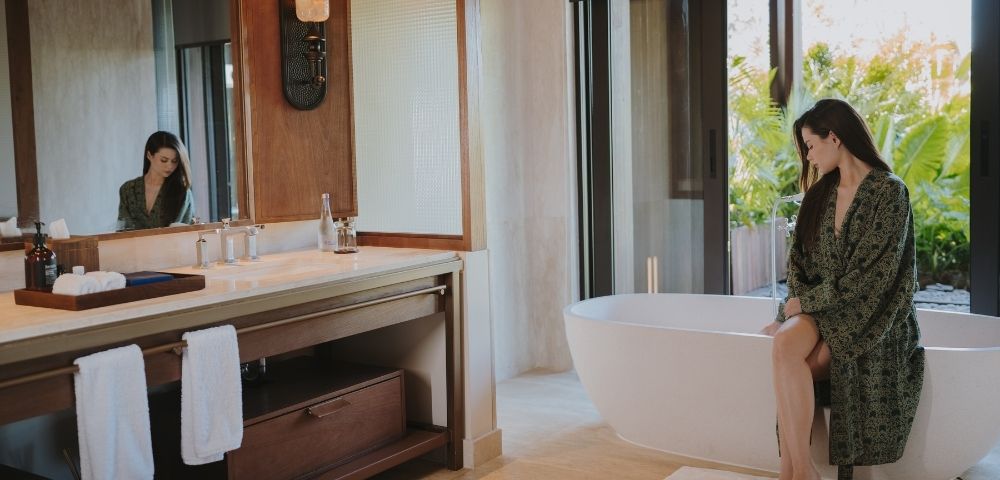 Woman in a green robe sitting on the edge of a white bathtub in a modern bathroom with large windows overlooking lush greenery.