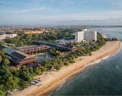 Aerial view of a sprawling resort complex with multiple low-rise buildings featuring dark, sloped roofs and a tall, white hotel building, situated alongside a sandy beach with lush tropical vegetation and the turquoise ocean.