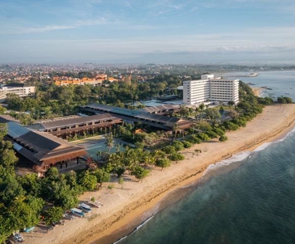 Aerial view of a sprawling resort complex with multiple low-rise buildings featuring dark, sloped roofs and a tall, white hotel building, situated alongside a sandy beach with lush tropical vegetation and the turquoise ocean.