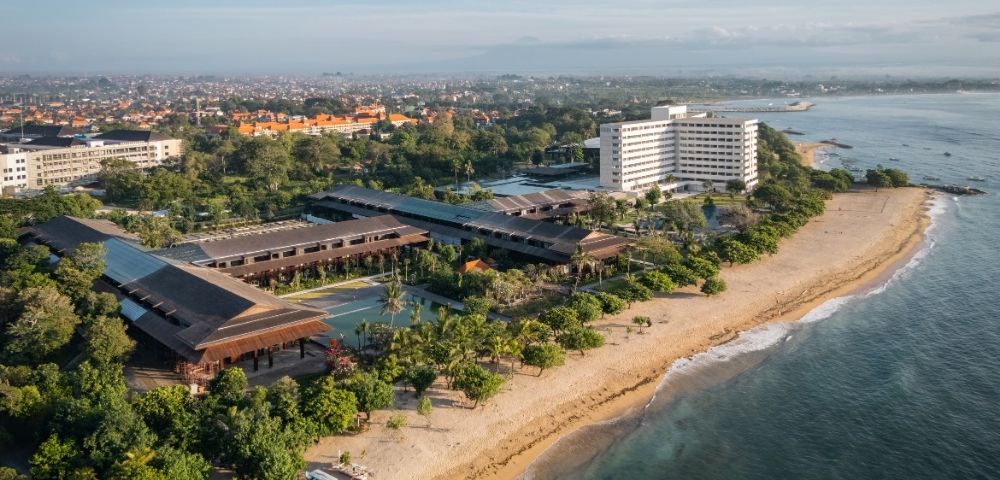 Aerial view of a sprawling resort complex with multiple low-rise buildings featuring dark, sloped roofs and a tall, white hotel building, situated alongside a sandy beach with lush tropical vegetation and the turquoise ocean.