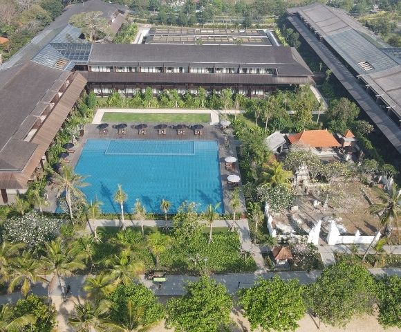 Aerial view of a large resort featuring a central outdoor pool, surrounding buildings, palm trees, and lush greenery.