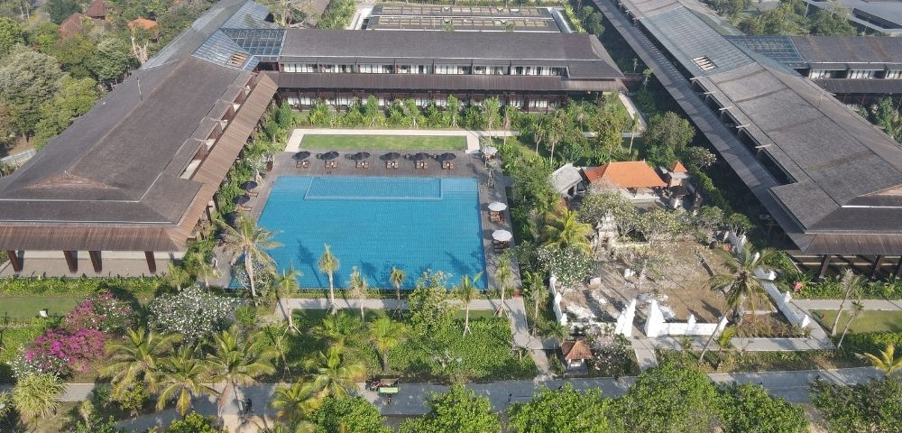 Aerial view of a large resort featuring a central outdoor pool, surrounding buildings, palm trees, and lush greenery.