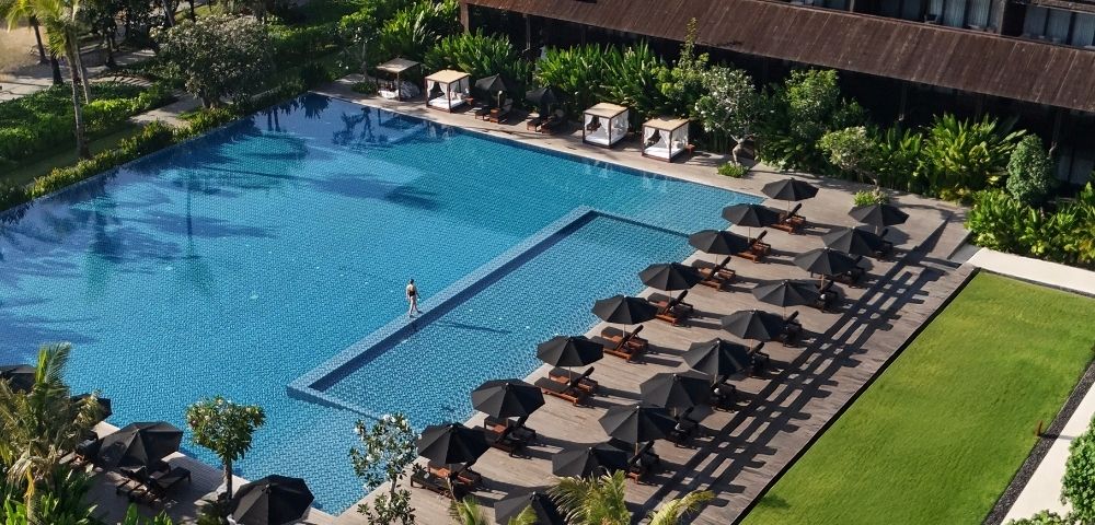 Aerial view of a large outdoor swimming pool with sun loungers and black umbrellas lined up on the deck beside a green lawn.
