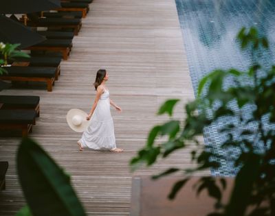 A woman in a white dress walks barefoot on a wooden deck by a pool, holding a sun hat. Lush green leaves frame the scene, conveying a serene, tropical vibe.