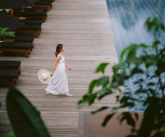A woman in a white dress walks barefoot on a wooden deck by a pool, holding a sun hat. Lush green leaves frame the scene, conveying a serene, tropical vibe.