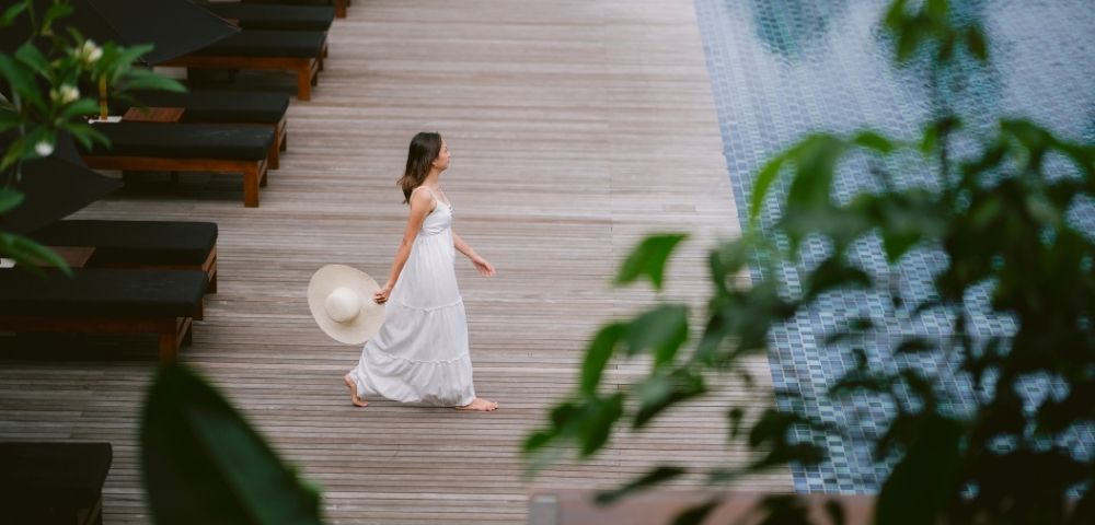 A woman in a white dress walks barefoot on a wooden deck by a pool, holding a sun hat. Lush green leaves frame the scene, conveying a serene, tropical vibe.