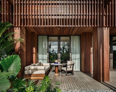A cozy patio features wicker furniture with beige cushions, a small round table, and lush greenery. Sunlight filters through wooden slats, creating a serene atmosphere.