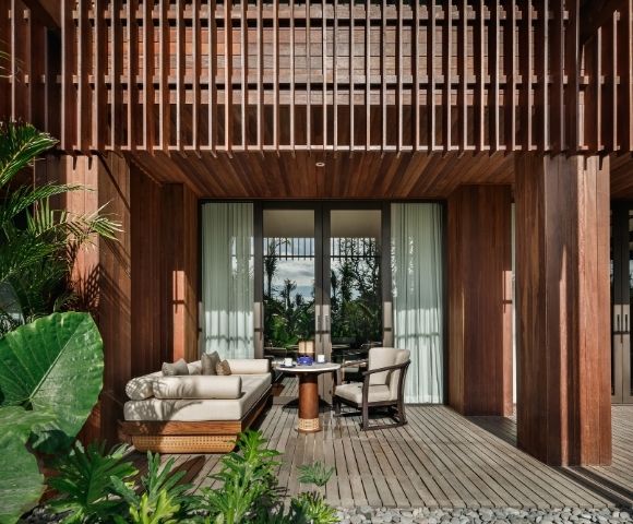 A cozy patio features wicker furniture with beige cushions, a small round table, and lush greenery. Sunlight filters through wooden slats, creating a serene atmosphere.