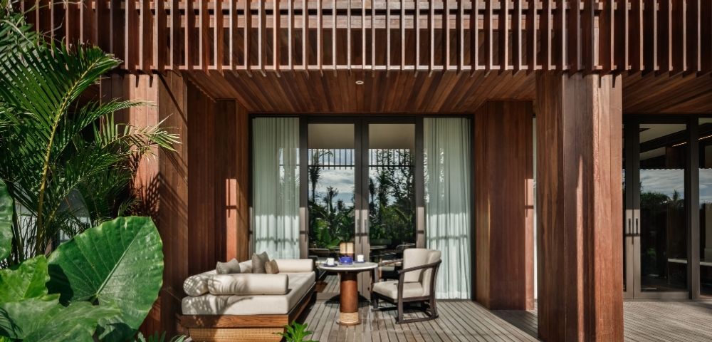 A cozy patio features wicker furniture with beige cushions, a small round table, and lush greenery. Sunlight filters through wooden slats, creating a serene atmosphere.