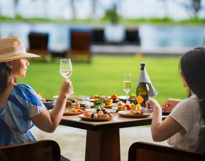 Two women enjoying a vibrant outdoor meal at a table laden with assorted dishes. One raises a glass of white wine, conveying a relaxed, joyful atmosphere.