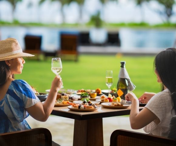 Two women enjoying a vibrant outdoor meal at a table laden with assorted dishes. One raises a glass of white wine, conveying a relaxed, joyful atmosphere.